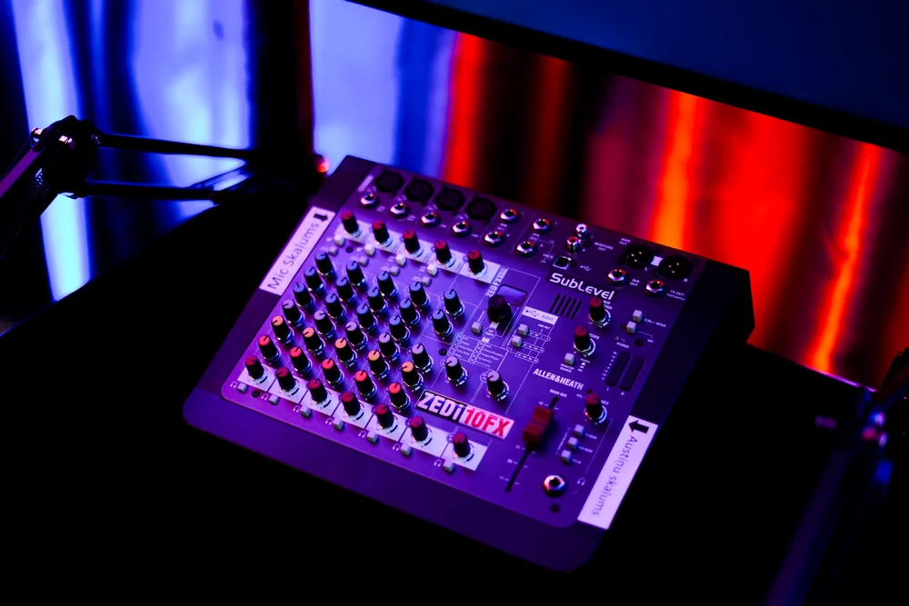 Tuvplāns ar Allen & Heath mikšerpulti podkāstu studijā, ar redzamām norādēm 'Mic Skaļums' un 'Austiņu skaļums' sarkanā un zilā apgaismojumā.